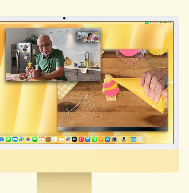 Gul iMac. Demonstration af Visning af skrivebord. Skærm viser en bager, der deltager i et videoopkald med en anden person. Den ene skærm viser bageren og mandens ansigt. Den anden skærm viser bagerens hænder, der pynter en småkage med glasur