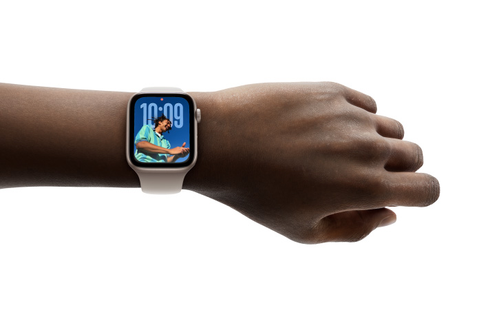 人の手首に装着された、ラバーバンド付きのローズゴールドのApple Watchのクローズアップ。新しいLiquid Glassのデザインの時刻アプリが表示されている