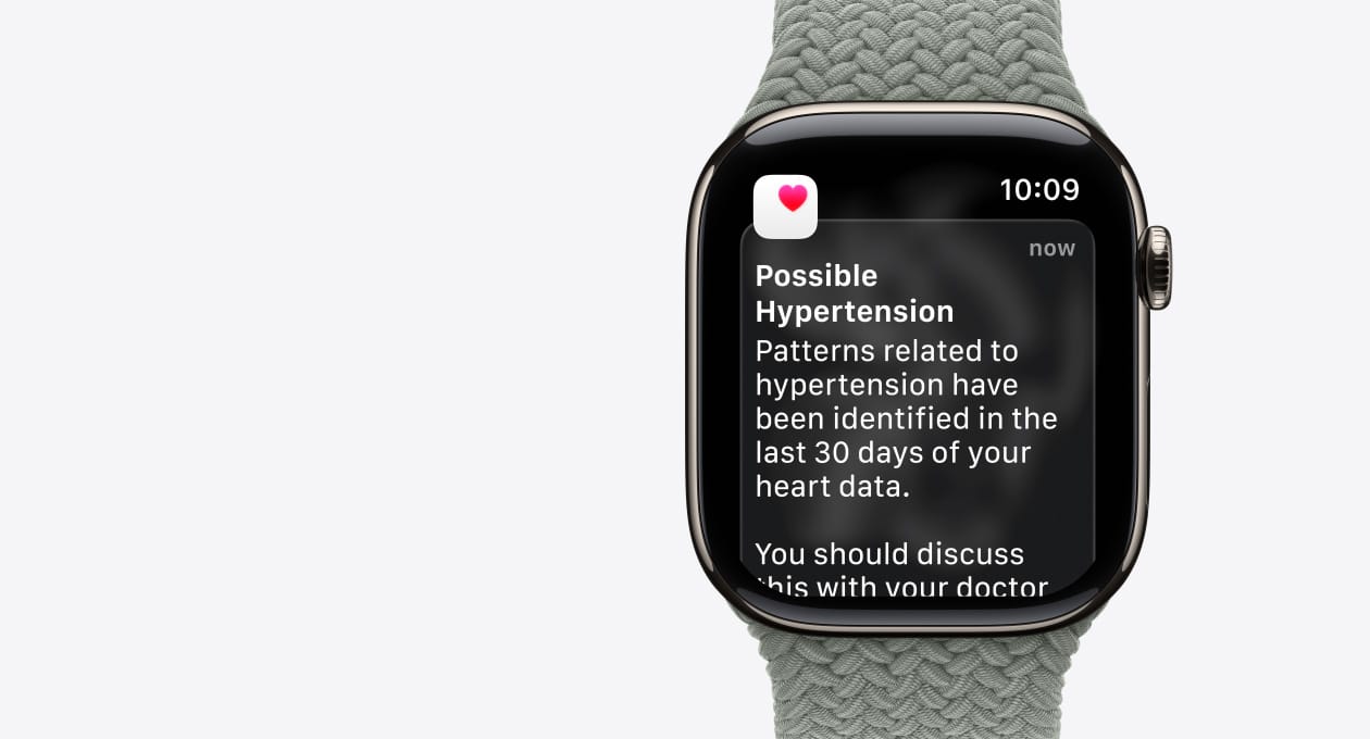 Apple Watch Series 11, gold colour, titanium case, possible hypertension notification, pink heart in white box, right side exterior, Digital Crown, strap, green grey Braided Solo Loop